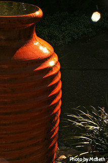 Self-contained ceramic water vase with contemporary (orange, brown) coloring beside tropical plant, large ceramic water fountains.