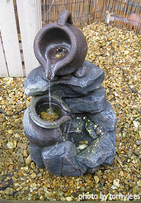 Small resin fountain shaped as tipped pot with spout pouring into lower pot with bamboo fence, miniature indoor/outdoor corner fountain, stone, rock look-alike fountain.