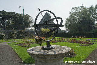 Metal sundial with arrow through middle mounted on stone pillar in garden, unique sundials, backyard garden decor.