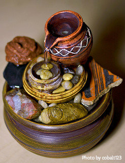 Small stone and pottery tabletop fountain (orange, yellow, brown, red) with small pot pouring into lower basin filled with decorative rocks, small decorative indoor fountain.